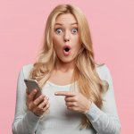 Blonde young female with pleasant appearance looks with terrified expression in smart phone, reads shocking news on webpage, isolated over pink background. Woman indicates at digital telephone
